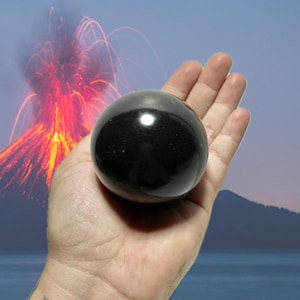 May include: A polished, black sphere held in an open hand. The sphere reflects light, creating a bright spot on its surface. The background features a volcano erupting with red and orange flames, and a blue sky.