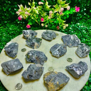 Puede incluir: Una colecci&oacute;n de 11 cristales de galena en bruto, grises y plateados, sobre una superficie de m&aacute;rmol blanco. Los cristales son de varios tama&ntilde;os y formas, algunos muestran un ligero brillo iridiscente.