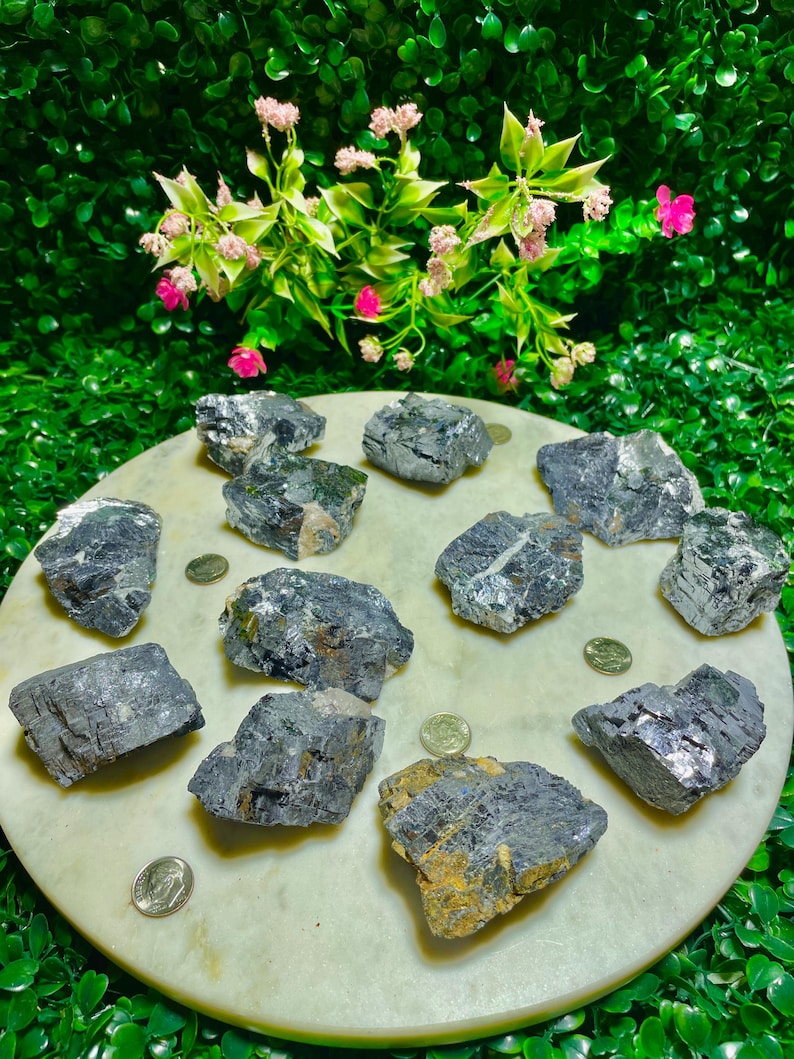 Puede incluir: Una colecci&oacute;n de 11 cristales de galena en bruto, grises y plateados, sobre una superficie de m&aacute;rmol blanco. Los cristales son de varios tama&ntilde;os y formas, algunos muestran un ligero brillo iridiscente.