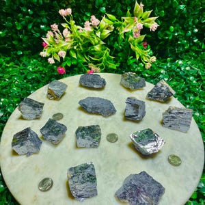 Puede incluir: Una colecci&oacute;n de 14 piedras crudas, grises, met&aacute;licas, con forma de cristal, sobre una superficie circular blanca. Las piedras son de varios tama&ntilde;os y formas.