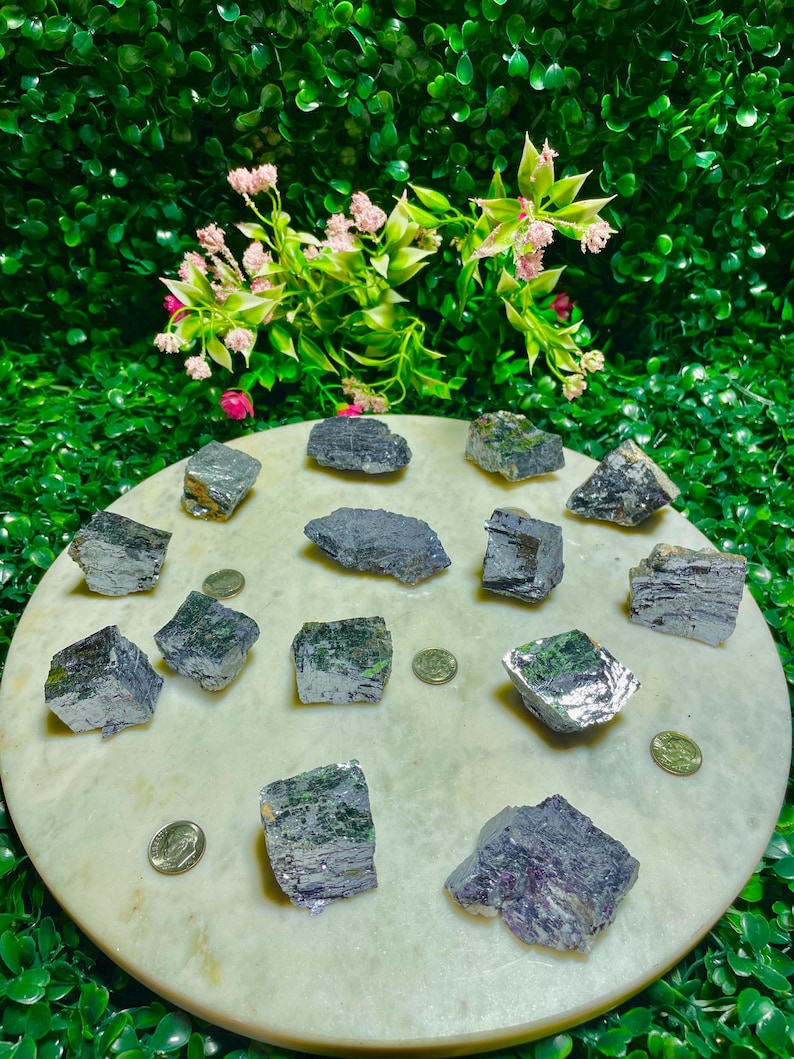 Puede incluir: Una colecci&oacute;n de 14 piedras crudas, grises, met&aacute;licas, con forma de cristal, sobre una superficie circular blanca. Las piedras son de varios tama&ntilde;os y formas.