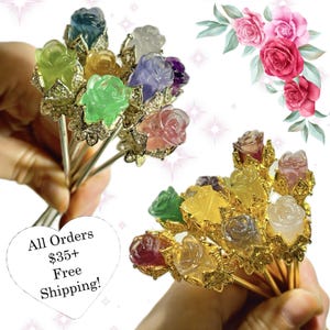 May include: A collection of hair sticks featuring rose-shaped gemstone heads in various colors, including blue, green, yellow, purple, pink, and clear. The roses are set on gold-colored metal stems with decorative filigree accents.