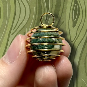 May include: A gold-colored wire-wrapped pendant holding a green and white stone. The pendant has a loop at the top for hanging. The stone has a marbled appearance with dark green and white patterns. The background is a green, abstract design.