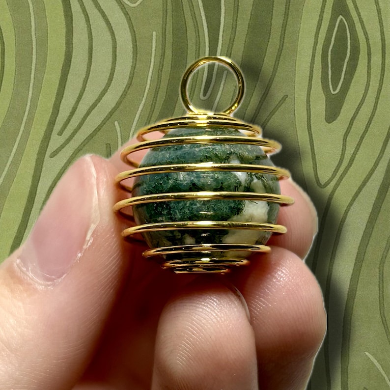 May include: A gold-colored wire-wrapped pendant holding a green and white stone. The pendant has a loop at the top for hanging. The stone has a marbled appearance with dark green and white patterns. The background is a green, abstract design.