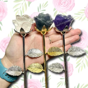 May include: Three gemstone rose sculptures in clear, gray, and purple. Each rose has a metallic stem and leaves. The clear rose has silver accents, the gray rose has gold accents, and the purple rose has silver accents. The roses are held in a hand.