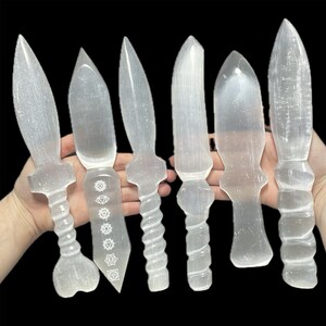 May include: Six selenite crystal daggers, some with etched chakra symbols, are displayed on a hand. The daggers are white and translucent.