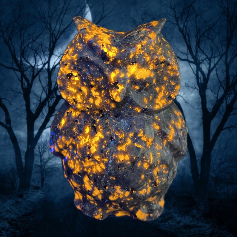K&ouml;nnte beinhalten: Eine geschnitzte Eulenfigur mit einem gesprenkelten Muster aus Orange und Dunkelblau. Die Eule ist vor einem dunkelblauen Hintergrund mit silhouettierten B&auml;umen und einem Mond platziert, was eine mystische Atmosph&auml;re schafft. Die Textur der Figur ist rau und nat&uuml;rlich.