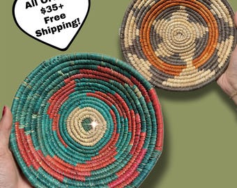 May include: Two woven, circular baskets. One is teal, red, and cream, with a spiral design. The other is brown, orange, and cream, with a starburst pattern. Both are held against a green background.