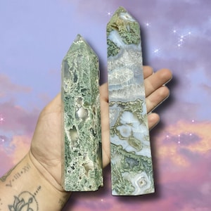 May include: Two green and white crystal obelisks. The obelisks have a natural, textured appearance with varying shades of green and white. One is a tall, rectangular shape, and the other is a shorter, more square shape. The background is a soft purple and blue.