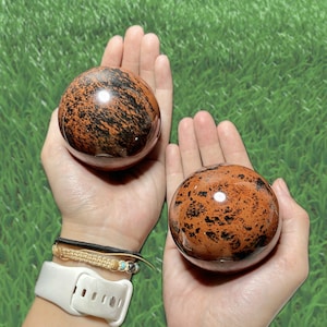May include: Two polished obsidian spheres, each held in a hand. The spheres are a mix of reddish-brown and black, with a glossy finish. The background is a field of green grass.