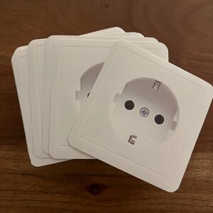 Könnte beinhalten: Fünf übereinander gestapelte weiße Kunststoff-Steckdosenabdeckungen mit einem Standard-Europastecker. Die Steckdose hat zwei runde Löcher und einen Erdungsstift.
