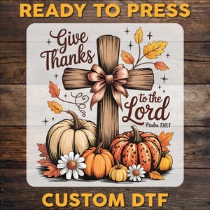 May include: A square DTF transfer featuring a wooden cross with a bow, surrounded by pumpkins, autumn leaves, and daisies. The text reads Give Thanks to the Lord Psalm 136:1. The design includes orange, brown, and white colors on a light background.