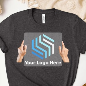 May include: A dark gray t-shirt with a clear sticker on the front. The sticker features a blue and white geometric design and the text "Your Logo Here".