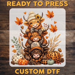 May include: A DTF transfer featuring three stacked Highland cows decorated with autumn elements. The top cow wears a knit hat with a sunflower, the middle has a leopard print bow, and the bottom wears a cap with the text FALL VIBES. Includes pumpkins, acorns, and orange leaves.