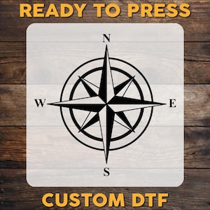 May include: Black compass rose design with the cardinal directions N, E, S, and W. The text "READY TO PRESS" is at the top, and "CUSTOM DTF" is at the bottom. The design is on a white background with a wood grain texture.