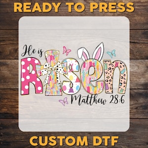 May include: A colorful Easter-themed design with the words "He is Risen" in a decorative font. The letter "R" is pink with white polka dots. The design includes butterflies, Easter eggs, and the text "Matthew 28:6". The words "READY TO PRESS" and "CUSTOM DTF" are also visible.
