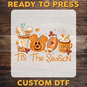 May include: A fall-themed design featuring a pumpkin with a Mickey Mouse face, a football, and a mug of hot chocolate. The words "Tis The Season" are written in orange script. The words "READY TO PRESS" and "CUSTOM DTF" are also visible.