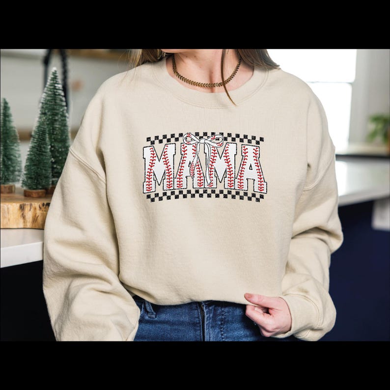 May include: A cream colored sweatshirt with a baseball-themed graphic that says "MAMA" in red and black. The graphic is surrounded by a black and white checkered border.