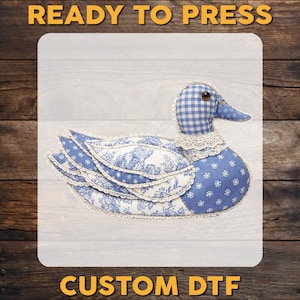 May include: A fabric duck with a blue and white floral pattern, a gingham head, and a lace collar. The duck has a blue beak and is labeled "READY TO PRESS" and "CUSTOM DTF" in gold text.