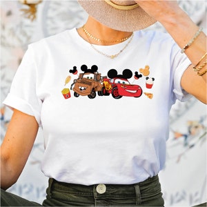 Pu&ograve; includere: Maglietta bianca con una grafica di Mater e Saetta McQueen di Cars, con orecchie di Topolino e icone di cibo Disney.