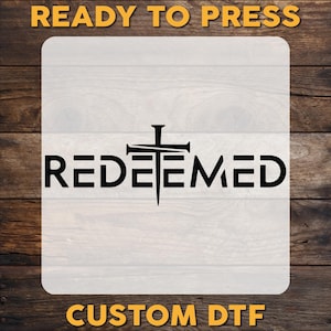 May include: A white iron-on transfer sheet with the word "REDEEMED" in black text and a black cross with three nails. The text "READY TO PRESS" is at the top of the sheet and "CUSTOM DTF" is at the bottom.
