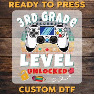 May include: A white controller graphic with the text "3rd Grade Level Unlocked" and the words "Ready to Press" and "Custom DTF". The design includes a red apple, a pencil, and a lock.