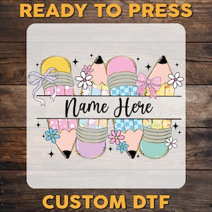 May include: A colorful design featuring pencils with bows and flowers, with the text "Name Here" in the center. The words "READY TO PRESS" and "CUSTOM DTF" are at the top and bottom.