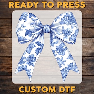 May include: A white bow with a blue floral pattern, ready to be pressed onto fabric using a heat press. The image is labeled "CUSTOM DTF".