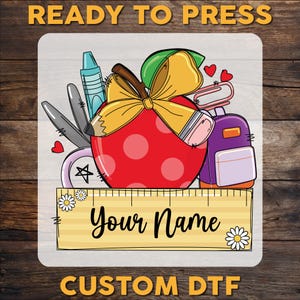 May include: A red apple with white polka dots and a yellow bow is surrounded by school supplies, including a pencil, scissors, a crayon, a book, and a purple backpack. The text "Your Name" is on a wooden ruler below the apple.  Custom DTF.