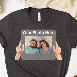 May include: A dark gray t-shirt with a white frame and the text "Your Photo Here" printed on the front. The frame contains a photo of a family of three, a man, a woman, and a child, smiling and lying on a wooden floor.