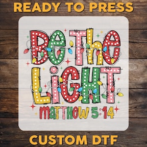 May include: A square DTF transfer sheet featuring the text BE THE LIGHT MATTHEW 5:14 in large, colorful, polka-dotted block letters. The design is wrapped in a string of multi-colored Christmas light bulbs and accented with small red star shapes.