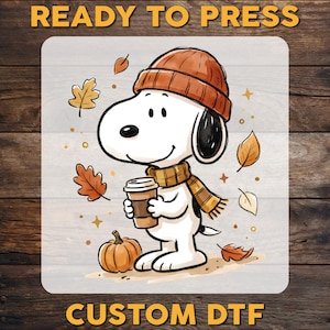 May include: A white cartoon dog wearing an orange beanie and a striped scarf, holding a brown coffee cup. The illustration features falling autumn leaves and a small orange pumpkin. The text READY TO PRESS and CUSTOM DTF appears in yellow.