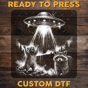 May include: Black and white graphic featuring a UFO beaming light onto two raccoons. One raccoon is standing with its arms raised, while the other raccoon is looking at the viewer. The words "READY TO PRESS" and "CUSTOM DTF" are at the top and bottom.