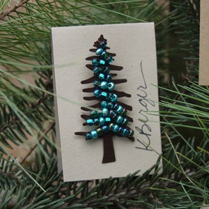 Puede incluir: Una tarjeta de papel marr&oacute;n con un recorte de un &aacute;rbol de Navidad decorado con cuentas iridiscentes verdes y azules. La tarjeta tiene la palabra "remarkable" escrita en ella.