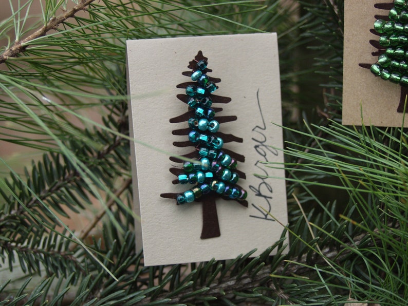 Puede incluir: Una tarjeta de papel marr&oacute;n con un recorte de un &aacute;rbol de Navidad decorado con cuentas iridiscentes verdes y azules. La tarjeta tiene la palabra "remarkable" escrita en ella.