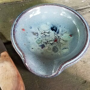 Op de afbeelding: Een blauwe keramische kaasschaal met een witte glazuur en rode en zwarte accenten. Naast de schaal ligt een kaasmes met een houten handvat.