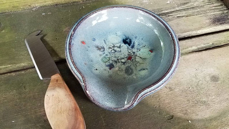 Op de afbeelding: Een blauwe keramische kaasschaal met een witte glazuur en rode en zwarte accenten. Naast de schaal ligt een kaasmes met een houten handvat.
