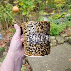 Könnte beinhalten: Eine braune und gelbe Keramik-Kaffeetasse mit einer strukturierten Oberfläche und dem Wort "Coffee" in Weiß geprägt.