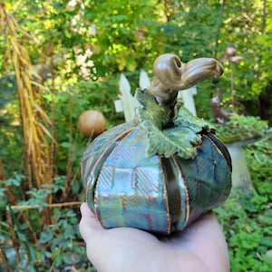 Puede incluir: Una calabaza de cerámica con un esmalte verde azulado y marrón, con una superficie texturizada. La calabaza tiene un tallo y hojas detallados, ideal para la decoración de otoño. El fondo es un exuberante jardín verde.