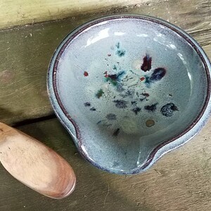 Op de afbeelding: Een blauwe keramische dipschaal met een gespikkelde glazuur en een bruine houten handgreep. De schaal staat op een houten oppervlak.
