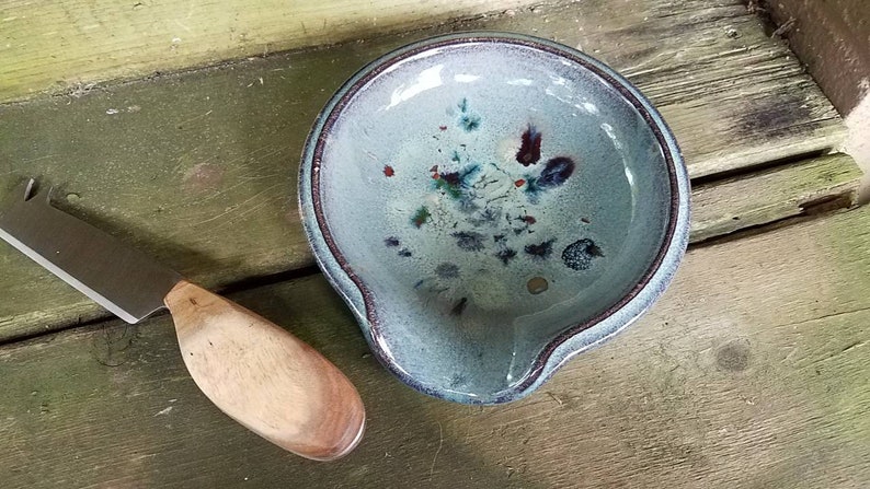 Op de afbeelding: Een blauwe keramische dipschaal met een gespikkelde glazuur en een bruine houten handgreep. De schaal staat op een houten oppervlak.