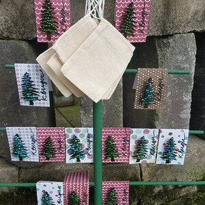 Puede incluir: Un soporte de metal verde con una base de madera presenta una variedad de peque&ntilde;os adornos de &aacute;rbol de Navidad hechos de cuentas. Los adornos se muestran en tarjetas rosas y blancas con un patr&oacute;n de copo de nieve u onda. Las tarjetas tienen la palabra "Kissed" escrita en ellas.