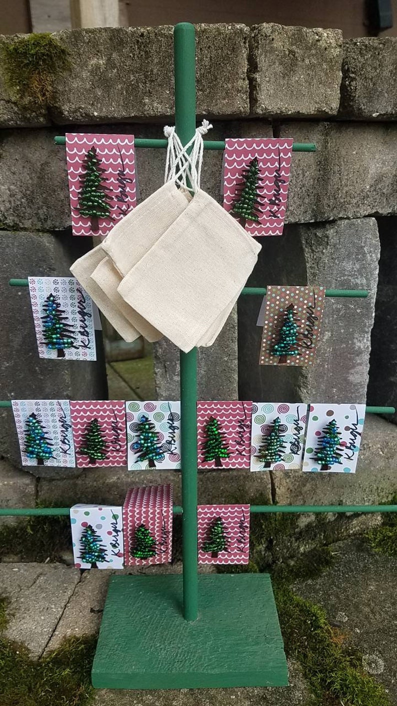 Puede incluir: Un soporte de metal verde con una base de madera presenta una variedad de peque&ntilde;os adornos de &aacute;rbol de Navidad hechos de cuentas. Los adornos se muestran en tarjetas rosas y blancas con un patr&oacute;n de copo de nieve u onda. Las tarjetas tienen la palabra "Kissed" escrita en ellas.