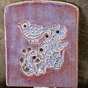 Puede incluir: Una luz de noche de cerámica con un diseño de pájaro y flores. La luz de noche de forma cuadrada presenta un fondo granate con un pájaro azul y un patrón floral. La luz de noche tiene una base blanca enchufable.