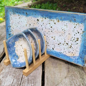 Könnte beinhalten: Ein rechteckiger Keramik-Servierteller und drei passende quadratische Teller mit einer gesprenkelten weißen und blauen Glasur. Die Teller werden in einem Holzständer ausgestellt. Der Teller und die Teller haben einen blauen Rand.
