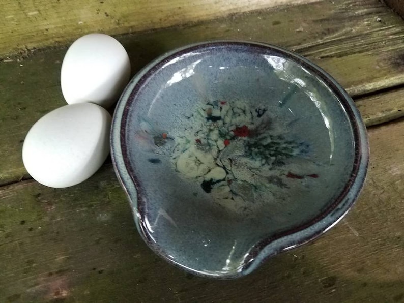Op de afbeelding: Een blauwe en witte keramische mengkom met een gespikkelde glazuur en een donkerpaarse rand. De kom heeft een diepe, ronde vorm met een brede, platte basis en een klein tuitje aan de ene kant.