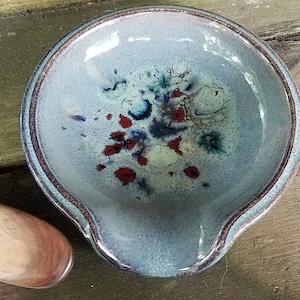 Op de afbeelding: Een blauwe keramische lepelsteun met een wit en rood splatterontwerp. De lepelsteun staat op een houten oppervlak.