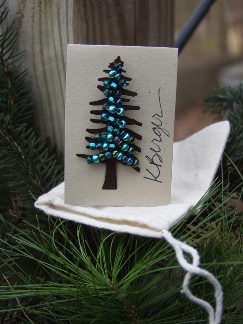 Puede incluir: Una peque&ntilde;a tarjeta blanca con una silueta de &aacute;rbol de papel marr&oacute;n decorada con cuentas verde azulado y verdes. La tarjeta tiene la palabra "Kreager" escrita en tinta negra.