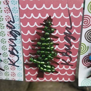 Puede incluir: Una tarjeta roja y blanca con un patr&oacute;n de ondas. La tarjeta tiene un &aacute;rbol de Navidad de cuentas verdes con la palabra "Navidad" escrita en negro.