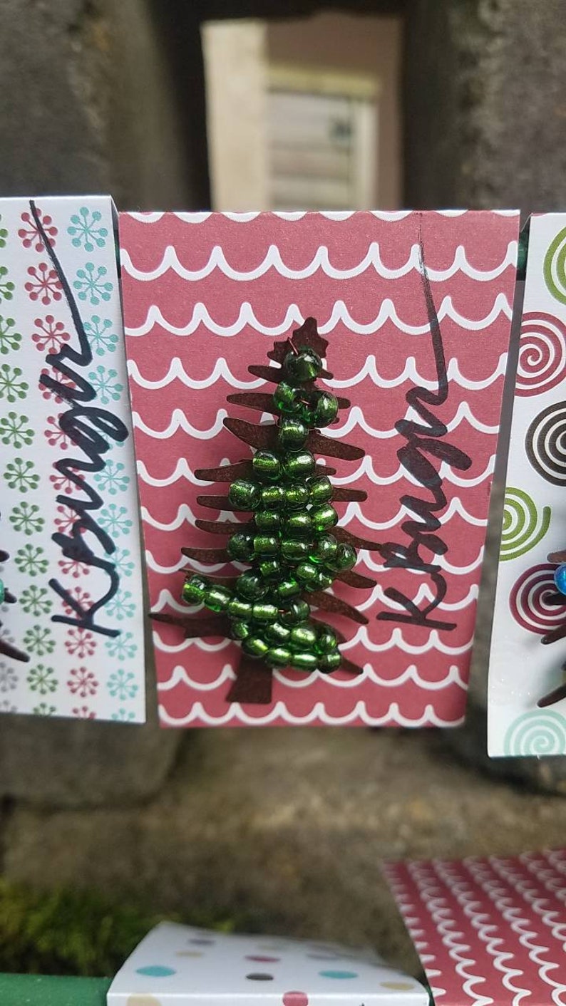Puede incluir: Una tarjeta roja y blanca con un patr&oacute;n de ondas. La tarjeta tiene un &aacute;rbol de Navidad de cuentas verdes con la palabra "Navidad" escrita en negro.